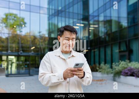 Asian man using phone outside office building, freelancer businessman typing message and smiling, programmer in casual clothes browsing online web pages from app. Stock Photo