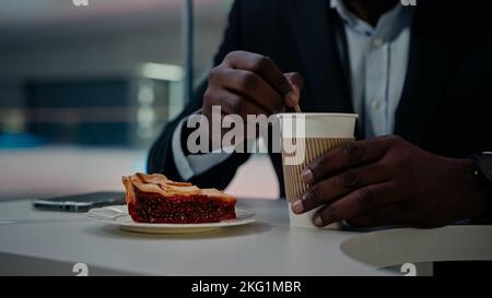 Unrecognizable African man worker businessman in formal suit sitting at table in cafe cafeteria restaurant stirs sugar in cup of aromatic coffee tea Stock Photo