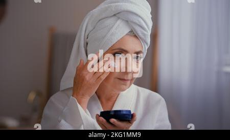 Portrait beautiful Caucasian old 60s woman 50s lady grandmother aged model with towel on head face care facial lifting hydration applies moisturizing Stock Photo