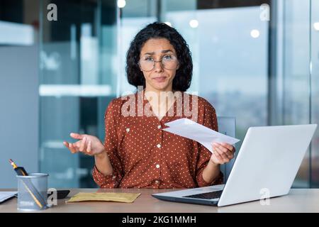 Portrait of disappointed business woman, latin american received notification letter from bank with bad news, office worker looking sad at camera, working at work using laptop. Stock Photo