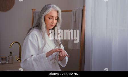Beautiful grandma 60s lady old senior gray haired female Caucasian woman in bedroom rubbing wrinkled hands applying natural hand cream moisturizing Stock Photo