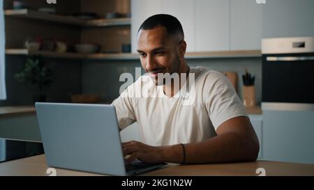 Smiling African millennial man guy American male user freelancer at home kitchen finish distant work with laptop satisfied with computer app online Stock Photo