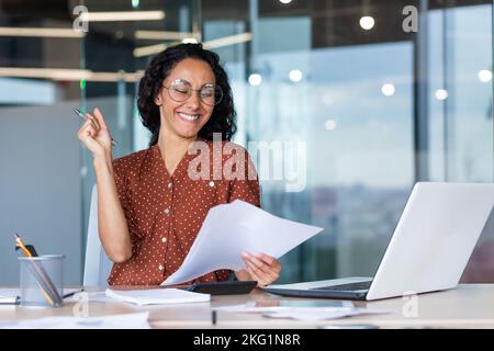 Businesswoman celebrating victory and successful achievement, Hispanic woman behind paper work reading documents with positive results reports, woman working on laptop inside office at table sitting. Stock Photo