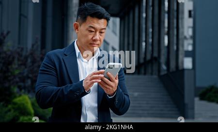 Asian businessman in city has mobile problem with telephone. Middle-aged CEO employer entrepreneur outdoors browsing phone feeling upset with broken Stock Photo