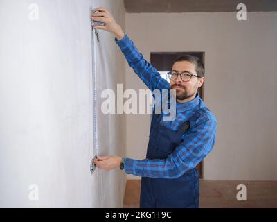 Side view of serious man using measuring tape in room with underfloor heating pipes. Designer in shirt taking measurements while working on renovation of apartment. Stock Photo