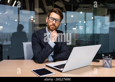 Serious thinking senior businessman working inside office using laptop, man in glasses solving problem, mature investor in business suit. Stock Photo