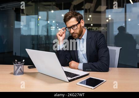Serious thinking senior businessman working inside office using laptop, man in glasses solving problem, mature investor in business suit. Stock Photo