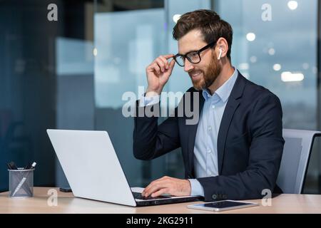 Mature businessman in headphones small earbuds talking on a video call using a laptop, boss at work at the desk in a business suit in the middle of the office smiling friendly. Stock Photo