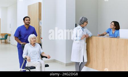 Healthcare assistant pushes wheelchair with senior woman in motion along corridor of hospital near reception desk with on-call nurse and doctor. Patie Stock Photo