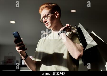 Side view portrait of Gen Z young man using smartphone while enjoying shopping at thrift store lit by sunlight Stock Photo