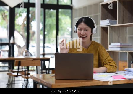 Friendly asian woman holds an online meeting while sitting in at home. Woman waving hand greeting participants of a video call. Online education Stock Photo