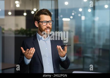 Friendly and smiling businessman in office recording audio message, man using smartphone app for communication, investor in business suit near window inside building. Stock Photo