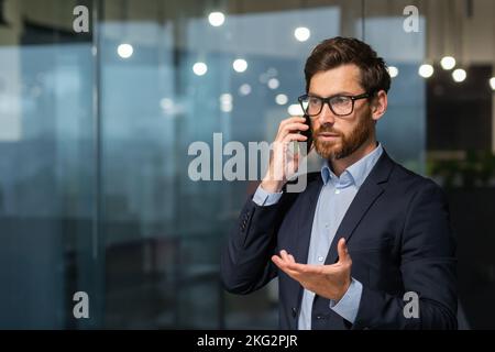 Serious and successful mature businessman inside office near window talking on phone, man in business suit and beard standing. Stock Photo