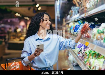 Woman shopper happy with phone in grocery department browsing shopping list in online app, choosing vegetables tomatoes in grocery department. Stock Photo