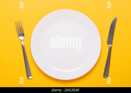 Clean empty white plate with knife and fork on yellow background place setting top view Stock Photo