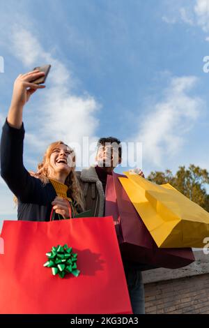 vertical photo of Multiracial couple of friends taking a very happy selfie holding shopping bags Stock Photo