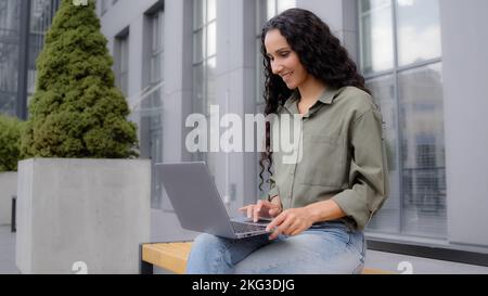Hispanic woman business lady 30s businesswoman freelancer sitting on bunch outdoors with laptop working in city online chatting typing computer Stock Photo