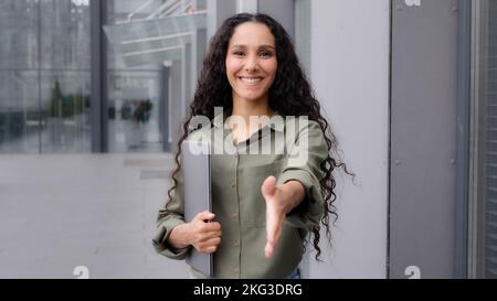 Caucasian 30s friendly businesswoman freelancer worker lady woman with laptop standing outdoors city holding hand in front welcome gesture hi sign Stock Photo