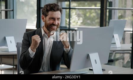 Exited happy Caucasian bearded businessman in office looking at computer with emotion winner cheerful rejoice with online win financial betting growth Stock Photo