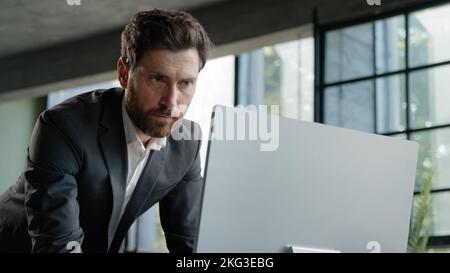 Caucasian busy bearded man adult 40s businessman employer standing in office has problem with paperwork typing data computer online check report Stock Photo