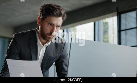 Serious 40s bearded businessman entrepreneur employee standing in office looking at papers report has problem with papers check data in online net Stock Photo