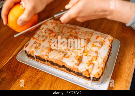 hands working with orange zest and grater over walnut cake with cream on wooden kitchen table Stock Photo