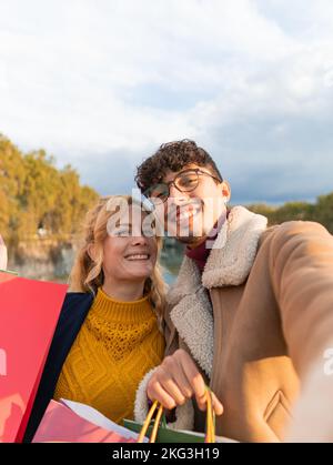Happy multiracial couple of friends taking a selfie in the city holding shopping bags in their hands Stock Photo