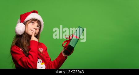A cute little Santa girl in a hat holds a New Year's gift in her hands, looks up dreamily and tries to guess the surprise in the box. Merry Christmas and Happy New Year. Isolated on green background. High quality photo Stock Photo