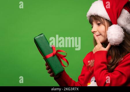 A cute little Santa girl in a hat holds a New Year's gift in her hands, looks at it dreamily and tries to guess the surprise in the box. Merry Christmas and Happy New Year. Isolated on green background. High quality photo Stock Photo