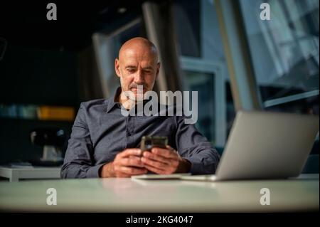 Mid adult business professional sitting at table with laptop and using mobile phone. Businessman text messaging and working in office. Stock Photo