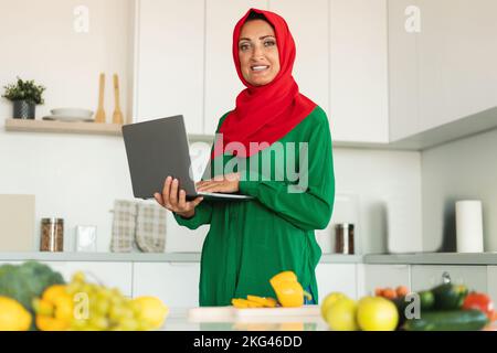 Searching for new recipes. Happy mature muslim woman holding and using laptop, cooking salad at home in modern kitchen Stock Photo