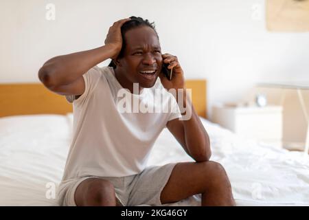 Oh No. Stresses Black Man Sitting On Bed And Talking On Cellphone Stock Photo