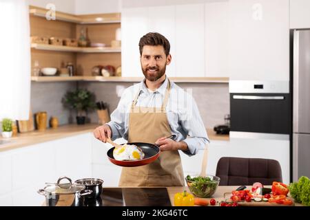 Smiling adult european bearded man chef in apron prepares scrambled eggs in modern kitchen interior Stock Photo