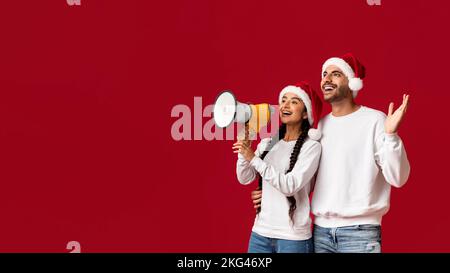 Arab Couple In Santa Hats Making Announcement With Loudspeaker Over Red Background Stock Photo