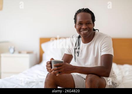 Handsome Black Man Resting On Bed With Cup Of Coffee In Hands Stock Photo