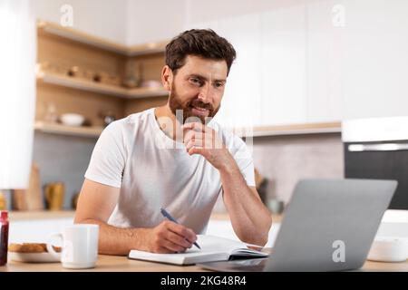 Happy handsome adult european bearded man making notes, watching video lesson on laptop Stock Photo