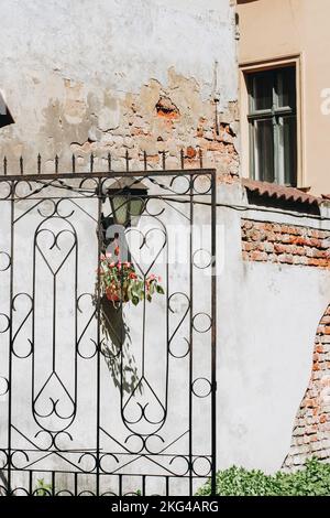 An old metal gate with a metal lantern hanging a flower pot with red flowers near a white building with crumbling red bricks Stock Photo