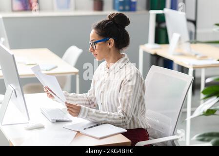 Focused businesswoman reading papers and business reports, sitting at workplace in modern office, free space Stock Photo