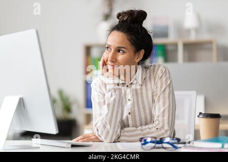 Dreamy young businesswoman thinking of new business strategy, sitting at workplace in office, looking aside and smiling Stock Photo