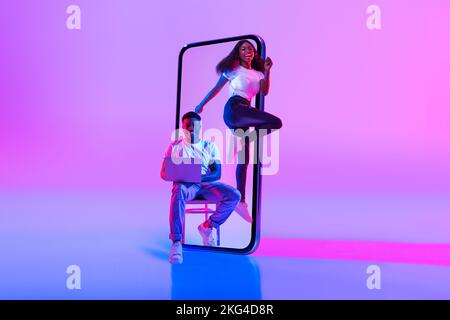 Cheerful excited young black lady jumping near huge smartphone enjoy freedom, man with laptop sits on chair Stock Photo