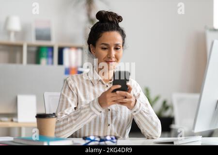 Smiling young female office worker using smartphone and reading message, sitting at table with pc in office interior Stock Photo