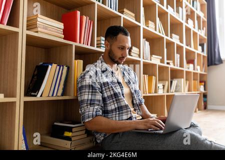 Young arab guy studying online from home, using laptop pc, sitting on floor near bookshelves, copy space Stock Photo