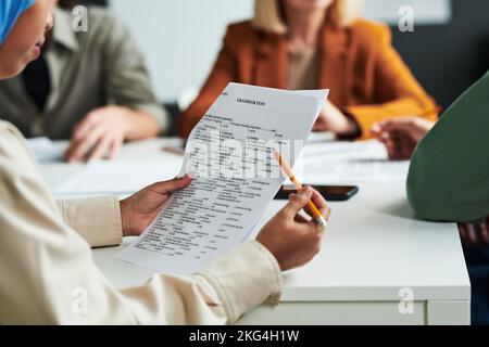 Selective focus on hands of Muslim female student with pencil looking through tasks of grammar test against teacher and young men Stock Photo