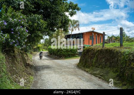 Filandia, Quindio, Colombia - June 6 2022: A Motorcycle Passing through a Dirt Road Surrounded by a lot of Trees, Plants and an Orange House Stock Photo