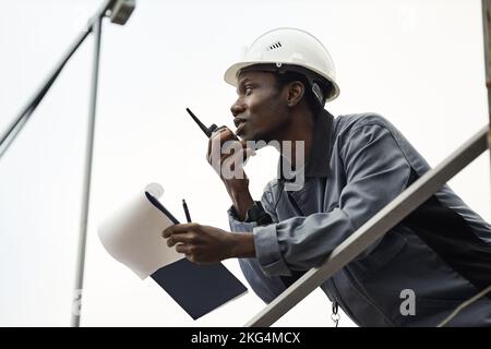 Graphic portrait of black male worker wearing hardhat and using radio against white sky Stock Photo