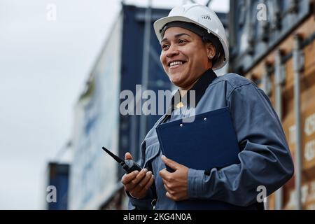 Waist up portrait of young black woman wearing hardhat and smiling while working at shipping docks with containers Stock Photo