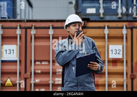 Waist up portrait of black woman wearing hardhat and talking to radio while working at shipping docks with containers Stock Photo