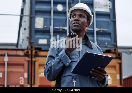 Waist up portrait of black male worker wearing hardhat and talking to radio in shipping docks with containers, copy space Stock Photo