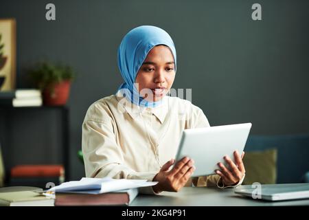 Young confident Muslim female tutor in earphones communicating with online audience while looking at screen of tablet during lesson Stock Photo
