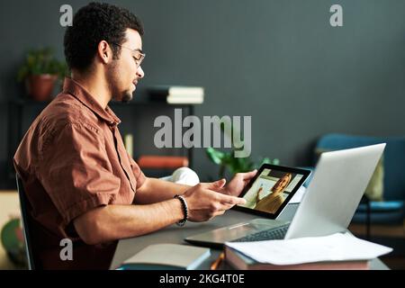 Young confident man looking at teacher making presentation on tablet screen and discussing points of new subject during online lesson Stock Photo